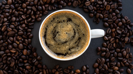 Top view of a cup of black coffee with crema, surrounded by roasted coffee beans on a dark background.