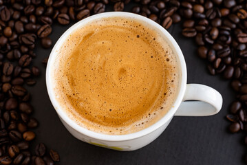 Top view of a cup of black coffee with crema, surrounded by roasted coffee beans on a dark background.