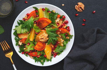 Autumn vegan persimmon salad with pomegranate seeds, walnuts, orange slices and lettuce, black background, top view