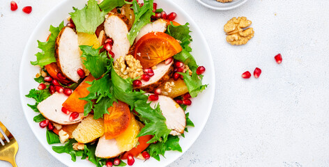 Autumn persimmon salad with grilled chicken fillet, pomegranate seeds, walnuts, oranges and lettuce, white table background, top view