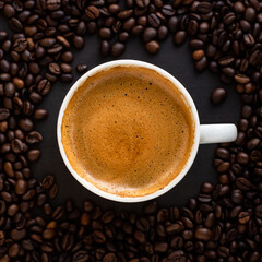 Top view of a cup of black coffee with crema, surrounded by roasted coffee beans on a dark background.