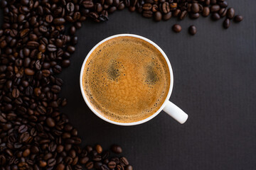 Top view of a cup of black coffee with crema, surrounded by roasted coffee beans on a dark background.