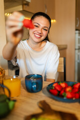 Smiling young woman showing a strawberry while having a healthy breakfast at home in her kitchen
