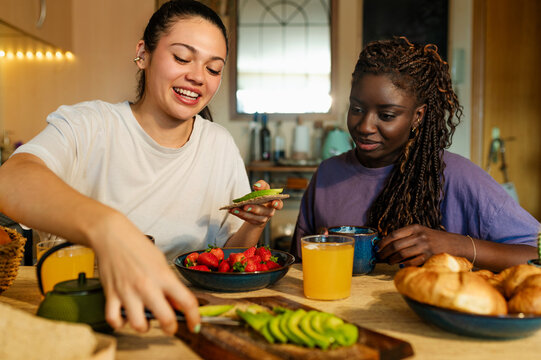 Two female roommates are having a healthy breakfast with fruit, avocado toast and orange juice in their home kitchen