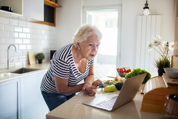Senior woman looking up recipes on laptop while preparing fresh vegetables in kitchen