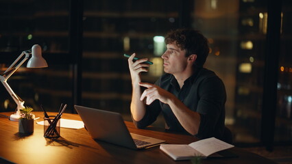 Night employee recording voice message by cellphone sitting dark office closeup.