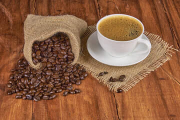 A jute bag with roasted coffee beans scattered on a rustic wooden table, next to it is a cup of freshly brewed coffee