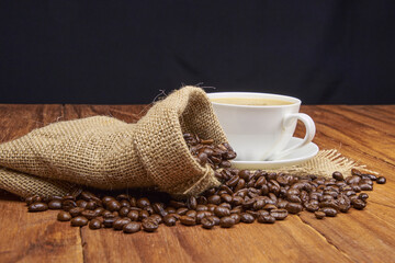 A jute bag with roasted coffee beans scattered on a rustic wooden table, next to it is a cup of...