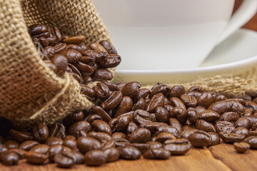 A jute bag with roasted coffee beans scattered on a rustic wooden table, next to it is a cup of...