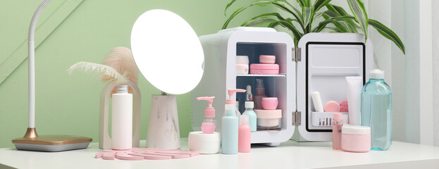 Small refrigerator with cosmetic products, lamp and mirror on table against green wall