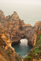 A natural sea arch is carved into the rugged golden cliffs of Ponta da Piedade, with a white haze...