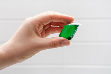 Female hand holding capsule with liquid washing powder on background and white tiles close-up.