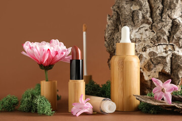 Concealer, green moss, flowers, lipstick, bottle of serum and tree bark on brown background, closeup
