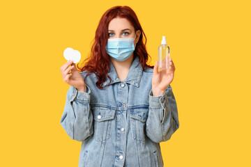 Beautiful young woman in mask with bottle of sanitizer and cotton pads on yellow background