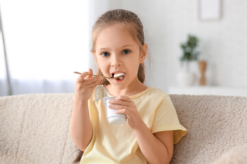 Cute little girl with spoon eating yogurt and sitting on sofa at home