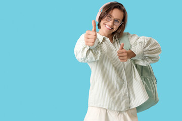 Happy female student with backpack and headphones showing thumb-up on blue background
