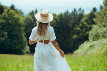 Blonde woman with straw hat and white dress walking through a green meadow on a sunny summer day