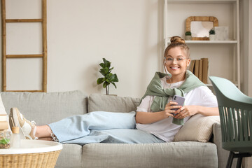 Young woman in eyeglasses lying on sofa and chatting with mobile phone in living room