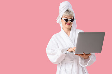 Portrait of beautiful young woman in bathrobe and sunglasses using laptop on pink background