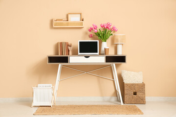 Stylish workspace with modern laptop, books and bouquet of beautiful tulip flowers near beige wall