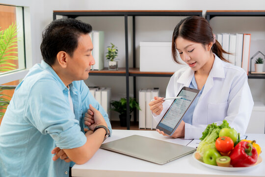 Asian nutritionist using tablet to explaining personalized meal plan with AI to patient during healthcare consultation, healthy lifestyle, woman dietitian showing nutrition plan with AI on tablet.