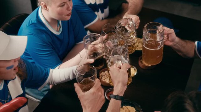 High angle of friendly football fans gathered by table in pub chatting about last match and clinking with glasses of beer over bowls with snacks while celebrating Club World Cup final