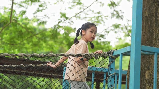 Happy young girl with pigtails waving cheerfully while standing on a net structure in a lush green outdoor setting surrounded by nature