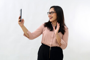 Portrait of friendly young Asian woman wearing glasses, smiling while having mobile conversation on video chat app, waving to smartphone camera, greeting someone, standing on white background.