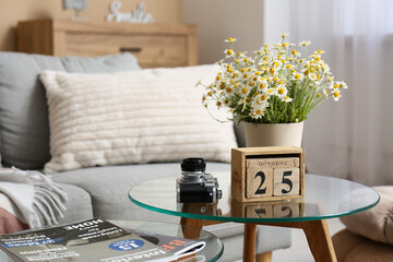 Cube calendar with date 25 OCTOBER, chamomiles and photo camera on table in living room