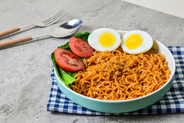 close up view of fried instant noodle with sliced of boiled chicken egg, tomato and mustard leaves on the plate. napkin. blue and white. grey or gray cement as background. fork, spoon.