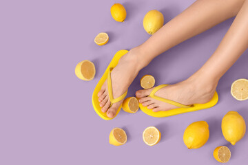 Female legs in yellow flip-flops with lemons on lilac background, top view