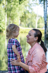 A joyful mom kneels to interact with her smiling daughter at a serene lake park. The sun shines...