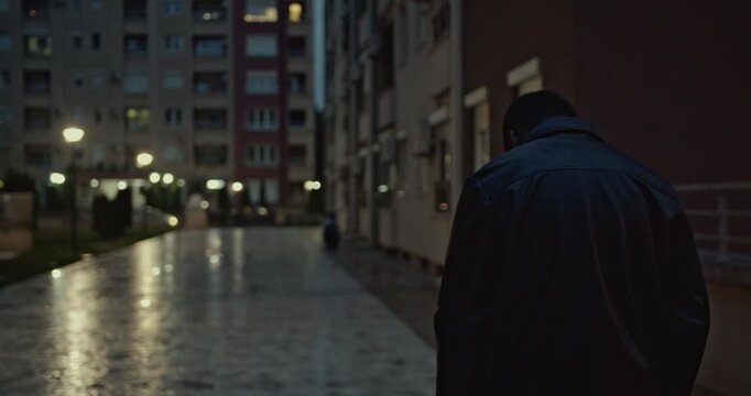 A young man walks through his neighborhood at night. He is wearing a leather jacket and appears to be lost in thought.