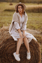 Front view of cheerful young woman sitting on haystack in field. Girl in beige dress, sweater on...