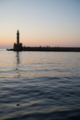 Silhouette of a lighthouse at sunset with calm water and distant figures on the horizon. The sky transitions from warm to cool tones, creating a serene and tranquil atmosphere.