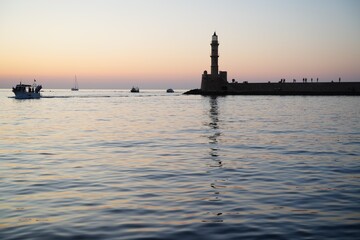 Lighthouse overlooking a peaceful harbor during sunset with boats sailing nearby. Silhouettes of...