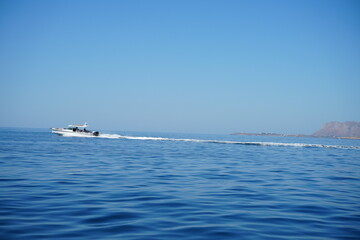 Naklejka premium Speedboat gliding across the expansive blue sea with distant rocky shoreline. Clear sky and tranquil water enhance the serene yet exhilarating scene of adventure and exploration.