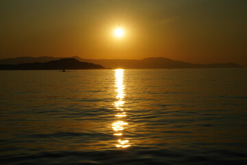 Naklejka premium Tranquil sunset scene with the sun setting over a calm sea, casting a golden reflection on the water. Silhouettes of distant mountains create a peaceful and serene ambiance