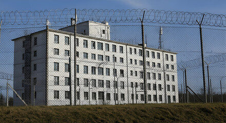 A large, imposing, white building with many windows is surrounded by a chain-link fence topped with barbed wire.