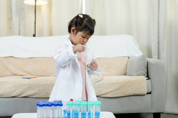Little Asian girl wearing white lab coat experimenting mixing liquids using pipette in colorful test tubes playful science education activity at home bright cozy living room early learning
