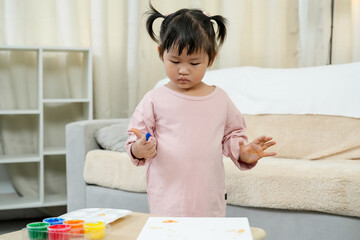 Little Asian girl wearing pink shirt holding paintbrush standing concentrating creating colorful...