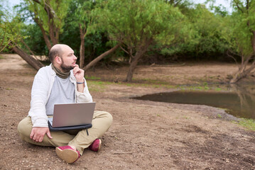 Man sitting outdoors, in nature, working on his laptop. He is thinking while contemplating the landscape. Concept: digital nomad working remotely while traveling, enjoying a lifestyle of freedom.