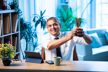 Mid age Indian businessman proudly holding trophy and smiling in stylish office