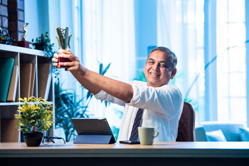 Mid age Indian businessman proudly holding trophy and smiling in stylish office