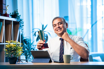 Mid age Indian businessman proudly holding trophy and smiling in stylish office