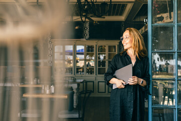 Happy Woman Holding Laptop Looking Outside Through Cafe Window, Wearing Smartwatch and Glasses