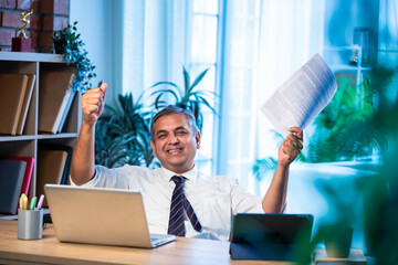 Indian professional shows triumph with papers in hand while seated at office desk