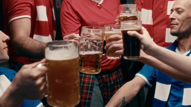 Tilt up of company of happy young male and female football fans wearing red and blue uniform meeting each other in pub, toasting with glasses of beer and discussing forthcoming match