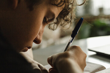 Close-up of teen boy writing in notebook with pen, focused face, studying at home indoors
