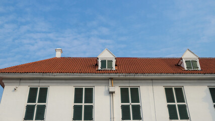 Colonial style white building with green shuttered windows and a red tiled roof under a clear blue...